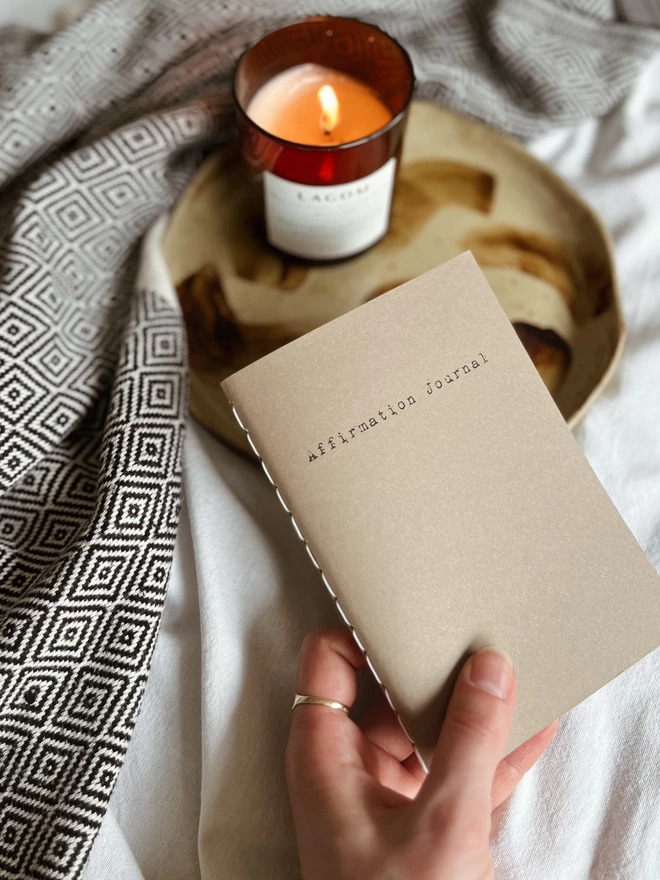 hand holding an a6 affirmation journal with a brown cover. A candle and blanket can be seen in the background