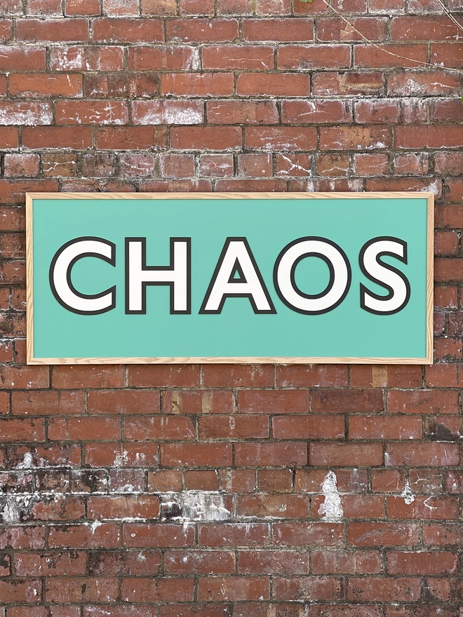 a wooden painted sign reading CHAOS on a aqua back board hung on a red brick wall