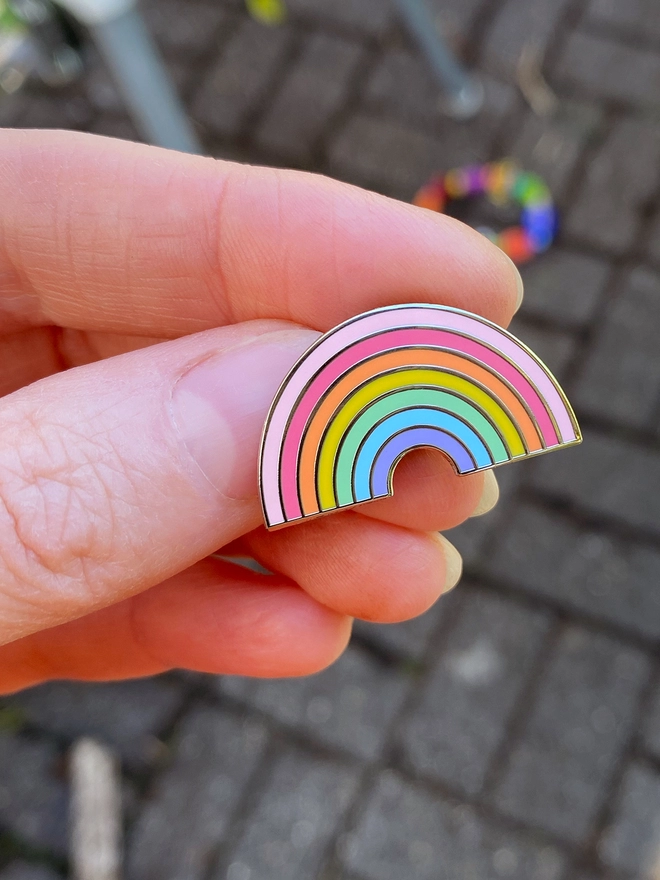 A small pastel rainbow enamel pin badge is being held.