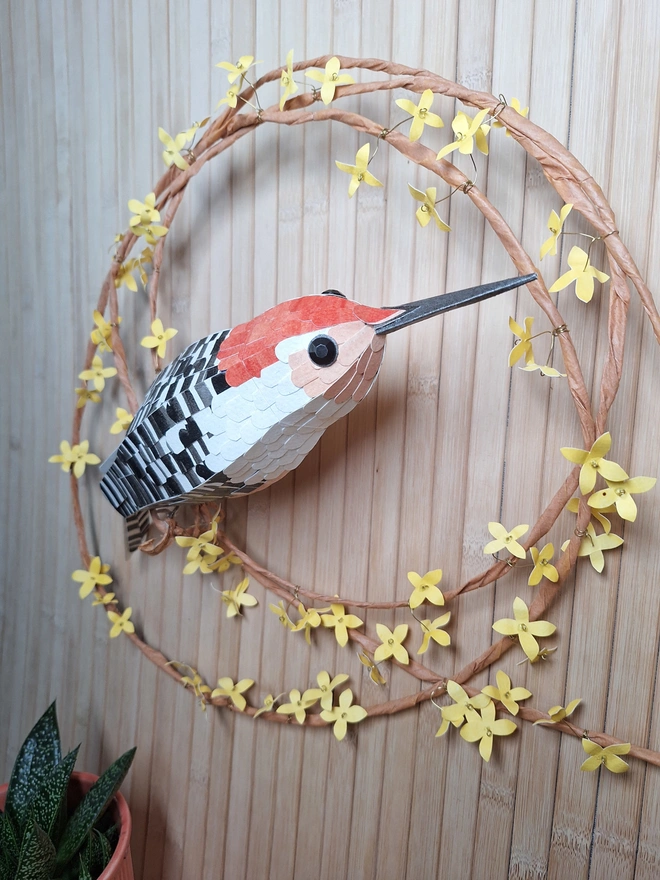Side close up of an original paper sculpture of an orange bellied woodpecker, perched on a wreath of yellow forsythia flowers