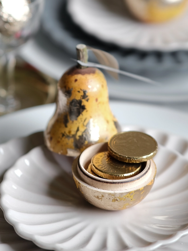Gilded vintage-style Christmas pear ornament opened to reveal a small surprise inside, shown on a festive Christmas table setting with white plates and ribbon.
