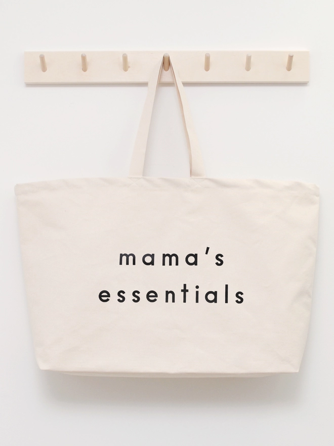 oversized natural canvas tote bag with mama's essentials slogan  hanging on a wooden hook