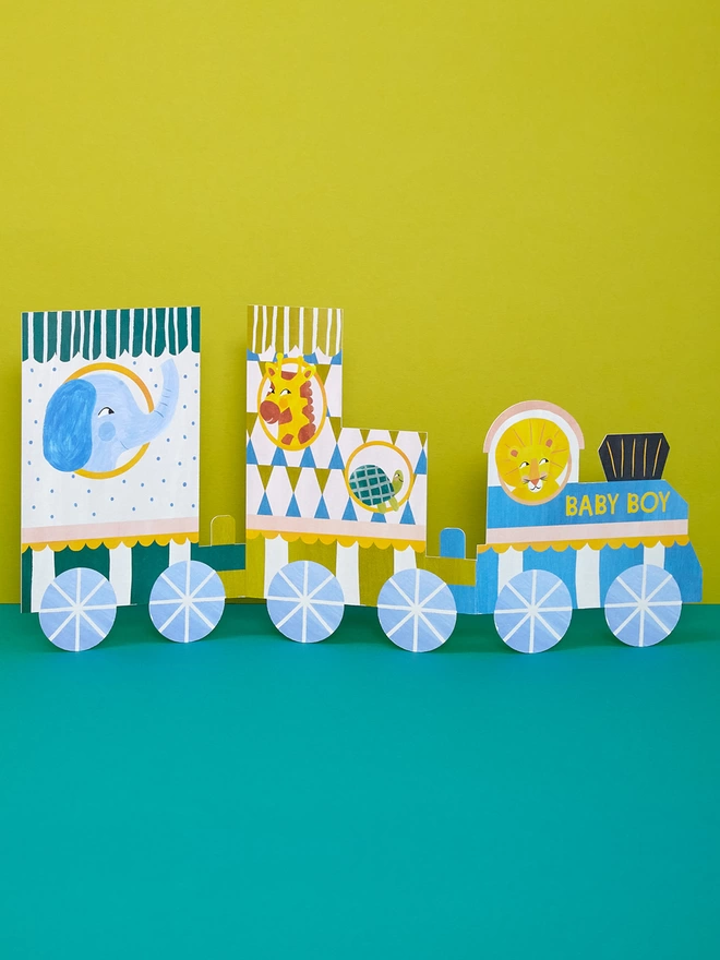 The concertina 'Baby Boy' fold-out card is a train with three carriages in vibrant blues, olives, greens and creams. Each carriage contains a character looking out the window, including an elephant, giraffe, tortoise and lion