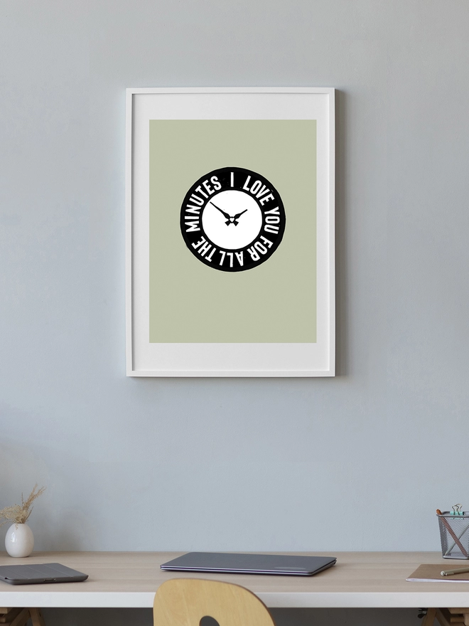 A framed green Woodism print hung on a wall in a home. The print features a black clock design with hand carved typography around it which reads: I Love You For All The Minutes