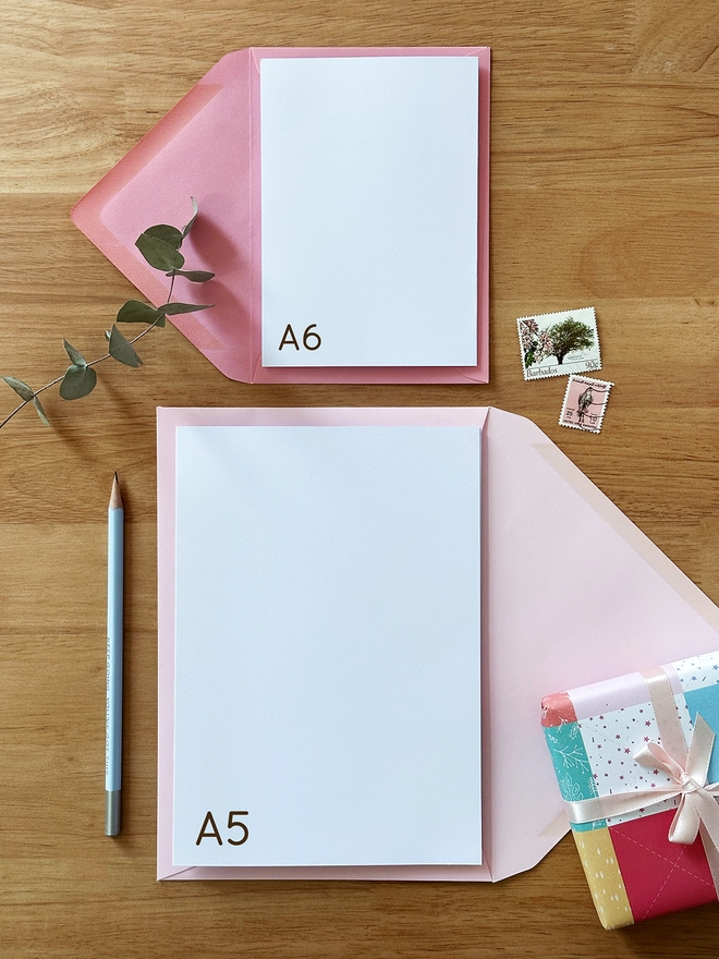 Two greetings cards with pink envelopes lay on a wooden desk, one is A6 size and one is A5 size.