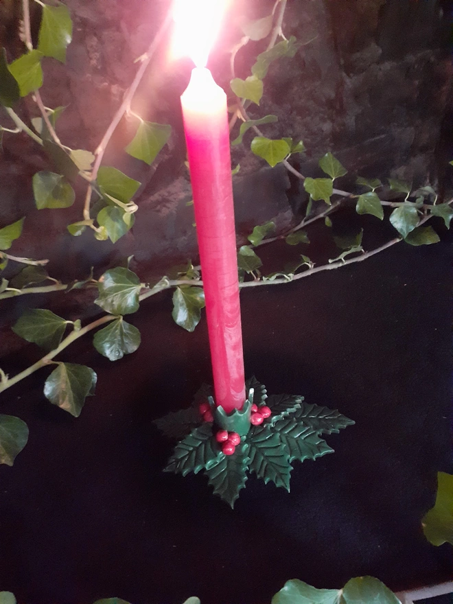 Tall red taper candle burning in a green holly leaf and red berry candle holder against a dark backdrop with ivy.