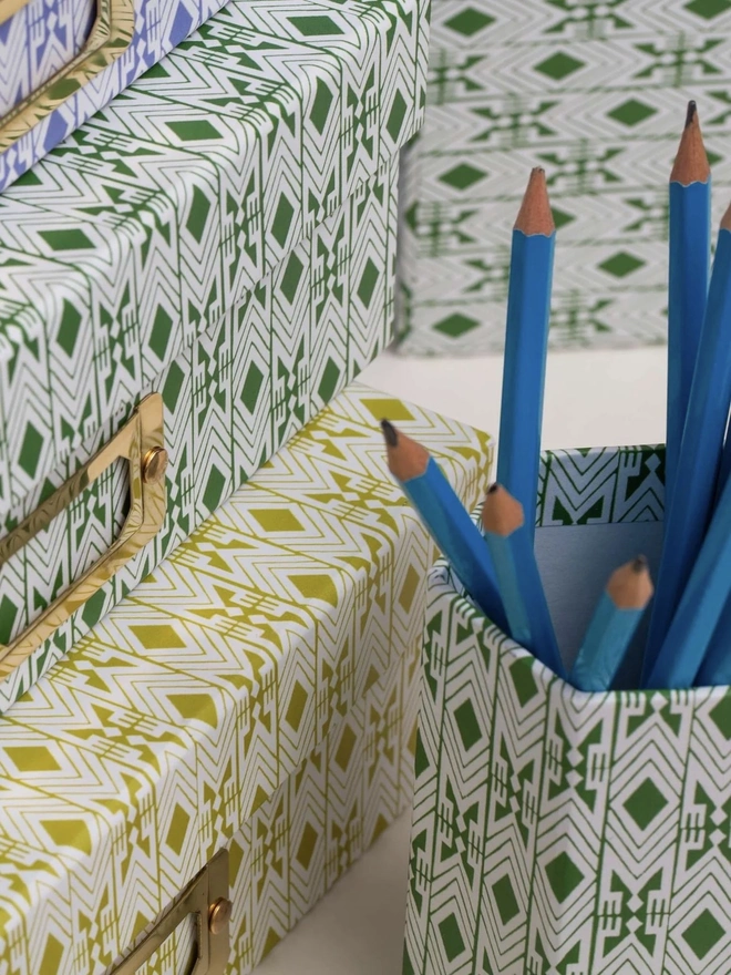 Tijan Hexagonal Brush Pot, a colourful patterned pencil pot sitting on a white surface accompanied by some folders. It is filled by some pencils. 