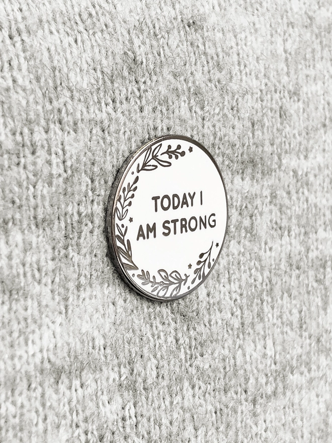 A round white enamel pin with a floral design and the words "Today I Am Strong" is pinned to grey fabric.