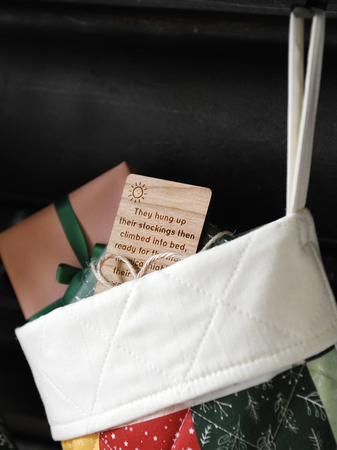 A patchwork Christmas stocking is hanging from a black mantlepiece. Presents and a wooden story card are peeking out of the top.