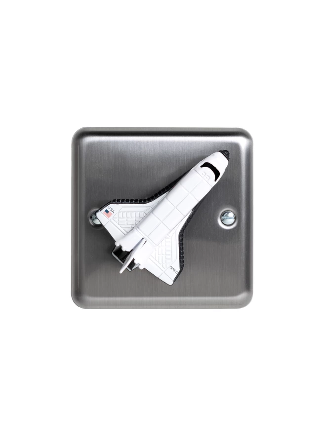 A brushed chrome dimmer light switch with a space shuttle as the rotary knob to turn the lights on and off on a white background. The children's light switch brand is Candy Queen Designs.