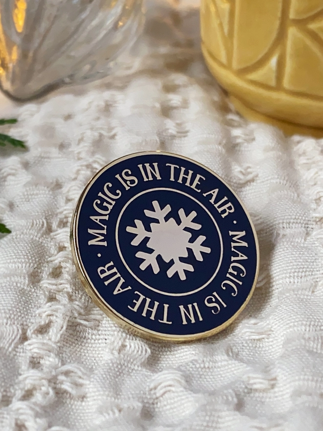 A navy blue and gold enamel pin badge with a gold star in the centre and the words "Magic is in the air" around the outside is on white fabric.