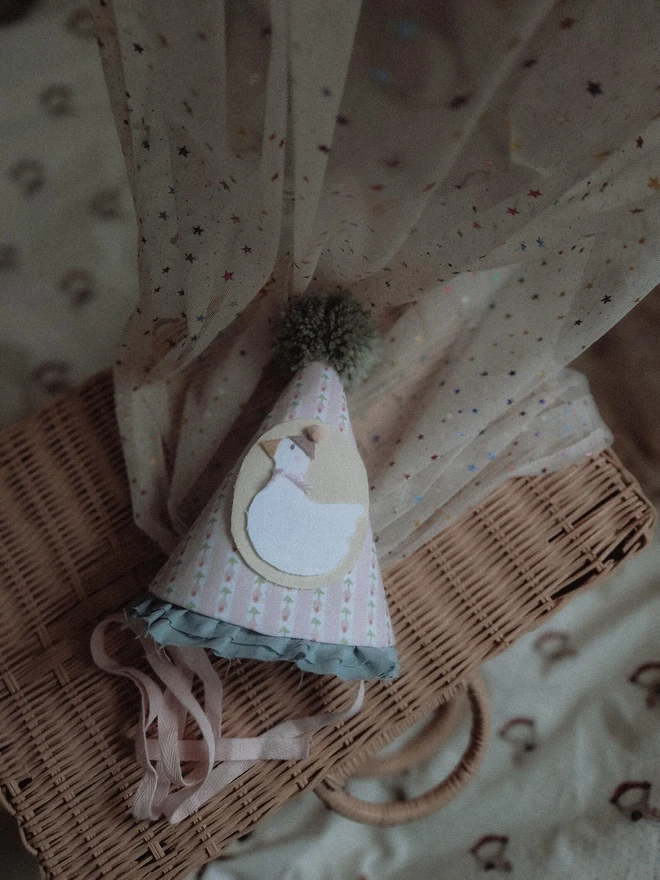 Baby Birthday Hat With Goose, a birthday party hat with a goose. It sits on a wooden hamper. 