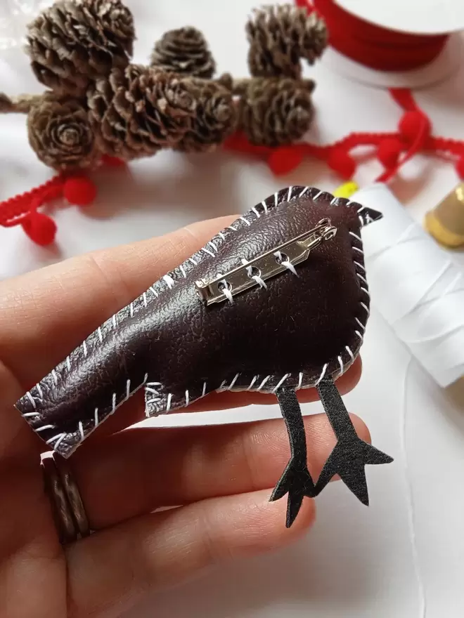 Showing the brown leatherette reverse side of a robin brooch, hand stitched around the edges, with brooch back, held in a hand amongst pine cones, red ribbon, pom pom trim, thimble and white reel of cotton