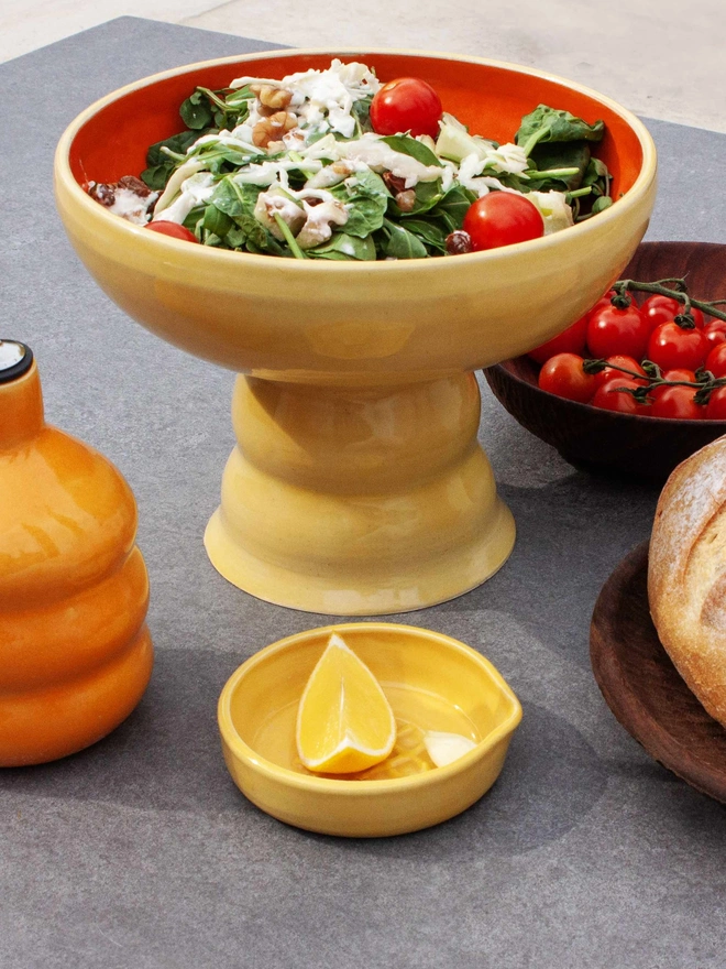 butter yellow pedestall bowl with a wide shallow bowl top and chubby foot. The bowl is yellow on the outside and orange on the inside. The bowl is filled with salad on a outdoor table surrounded by an oil bottle, lemon in a bowl, bread and tomatoes