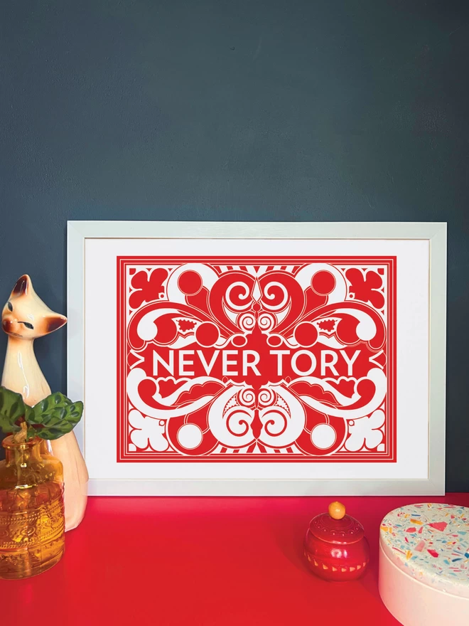 A bold, landscape red and white symmetrical illustration with Never Tory written in white at the centre. The print is in a white frame on a red cabinet, and is resting against a dark grey wall. Next to the frame is a cat ornament, an orange vase with a plant, a small red wooden pot and a white and multi coloured terrazzo pot.