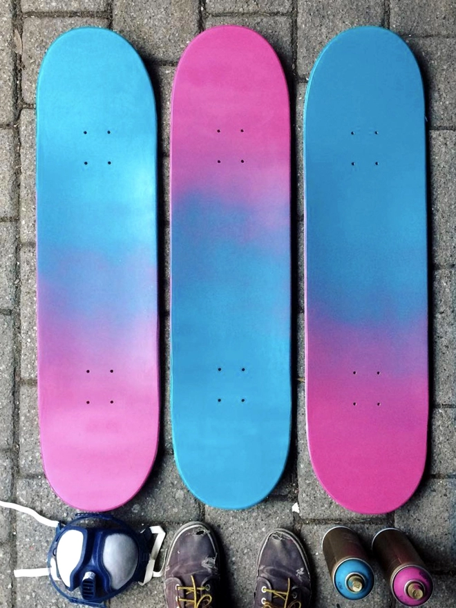 3 skateboards spray painted with a pink and blue blend / gradient lie on the floor alongside spray paint cans, a mask and the artist’s shoes. 