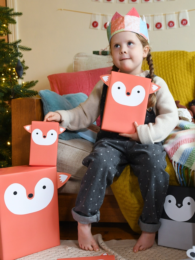A young girl wearing dungarees and a patchwork crown sits on a sofa beside a Christmas Tree and holds a gift wrapped as an orange fox.