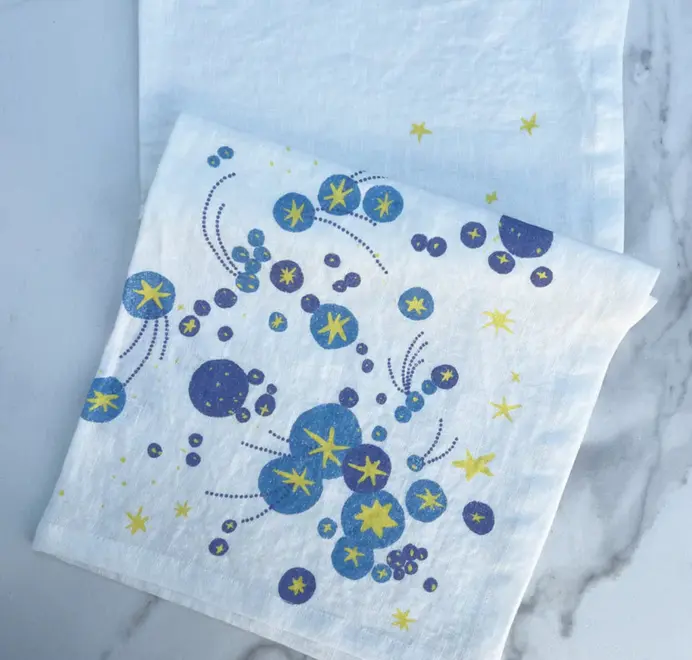 Solaris Napkins In White Or Blue. Off-white cloth napkin with blue and yellow starburst and orbital patterns on a white tablecloth. 