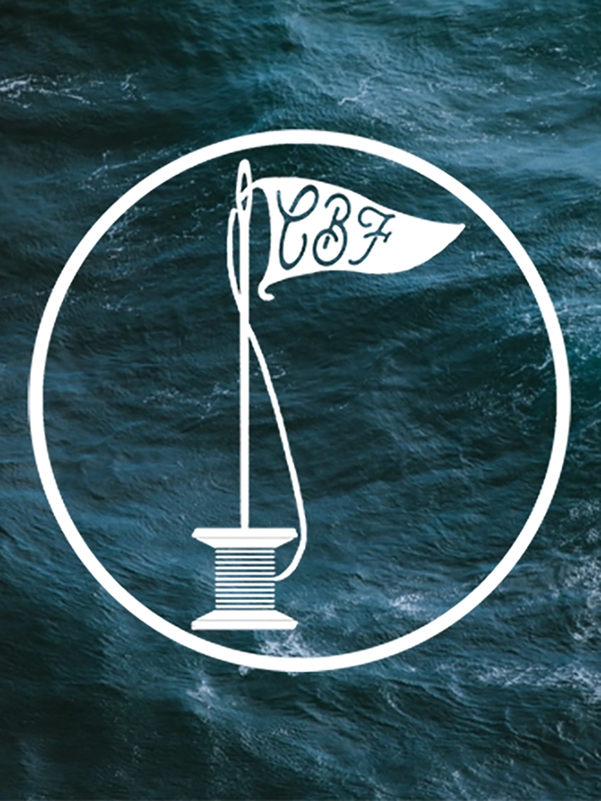 Caro B Fin Studio’s logo of a cotton reel with a needle in the top acting as a flag pole with a thread coming off the reel forming a flying pennant. It is on a background of a rough teal sea.