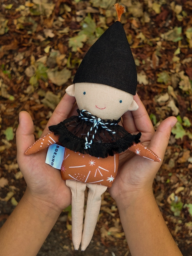 Handmade autumn elf doll with a black pointed hat, orange patterned top, black ruffled collar with black-and-white twine tie, embroidered blue eyes and pink smile, and light coloured face and legs 