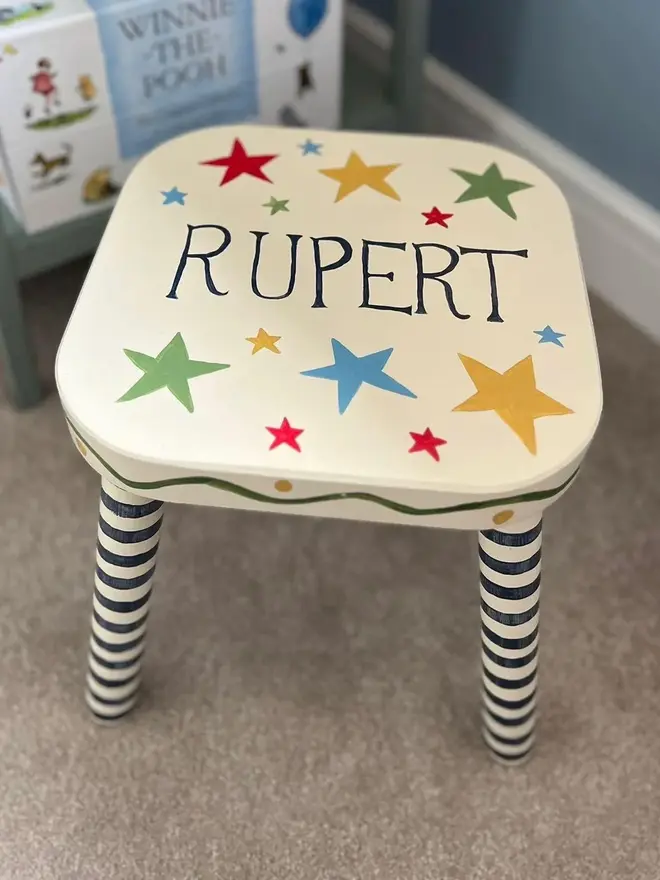 personalised hand-painted nursery stool. a small white stool with blue and white striped legs, painted with 'rupert' on top.