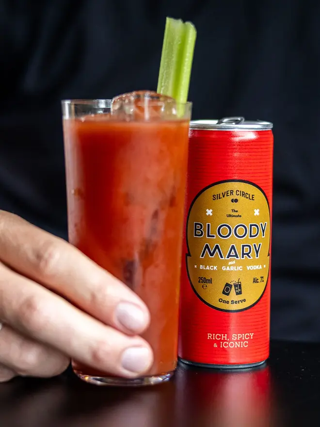 Ultimate Bloody Mary In A Single Serve Can, can of bloody mary next to a glass of bloody mary with a celery in it on a black surface. 