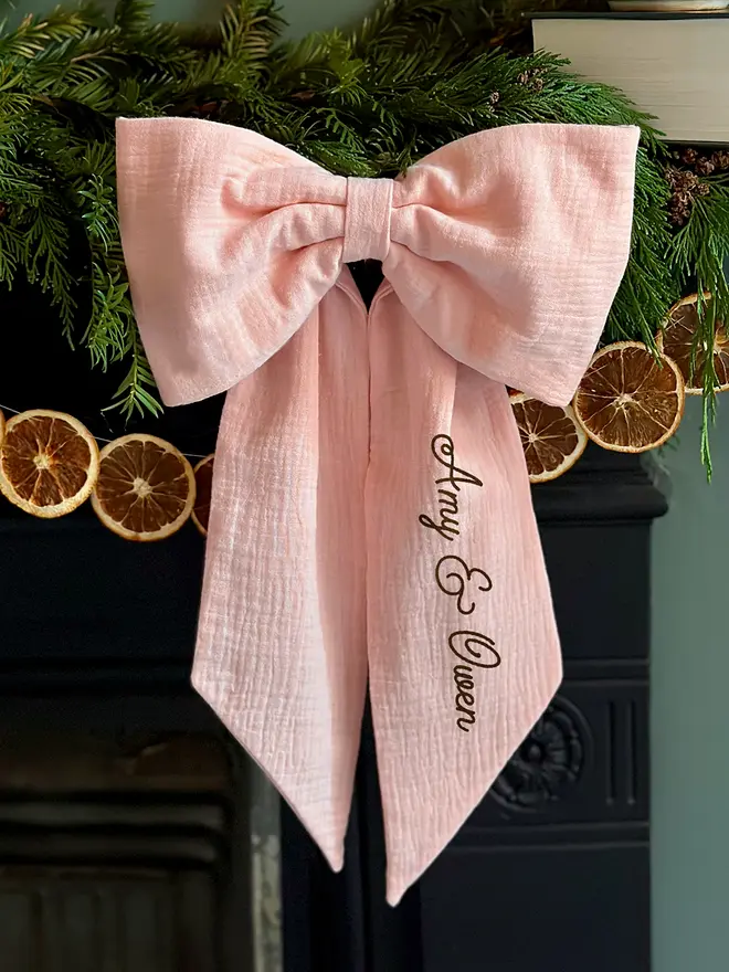 A handmade Christmas bow made from blush pink cotton with the words "Amy & Owen" on one tail is hung from a black mantlepiece that is decorated for Christmas with greenery, orange slice garland, and candles.