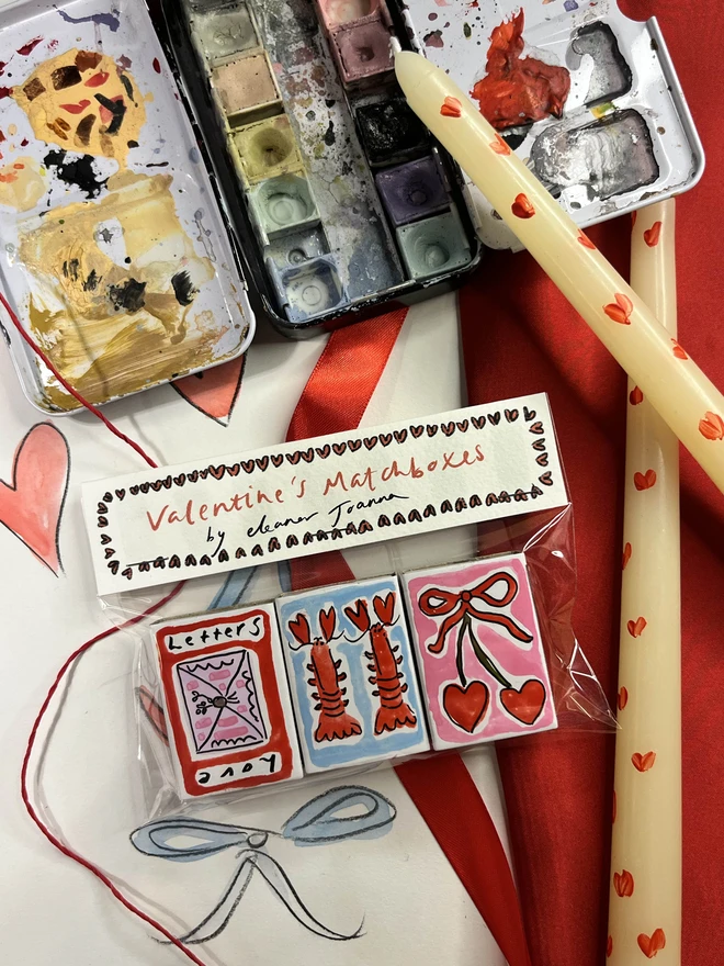 A collection of three hand-painted matchboxes with playful and colorful designs: one features two bright red lobsters, another displays vibrant cherries, and the third showcases a love letter. Each matchbox is crafted with whimsical illustrations and creative lettering, making them a delightful addition to any collection.