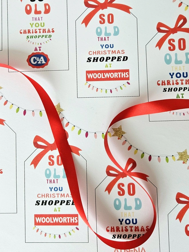 A close up photograph of the wrapping paper with tags that say so old you Christmas shopped at Woolworths, Tie Rack, C&A, Athena with a red ribbon.