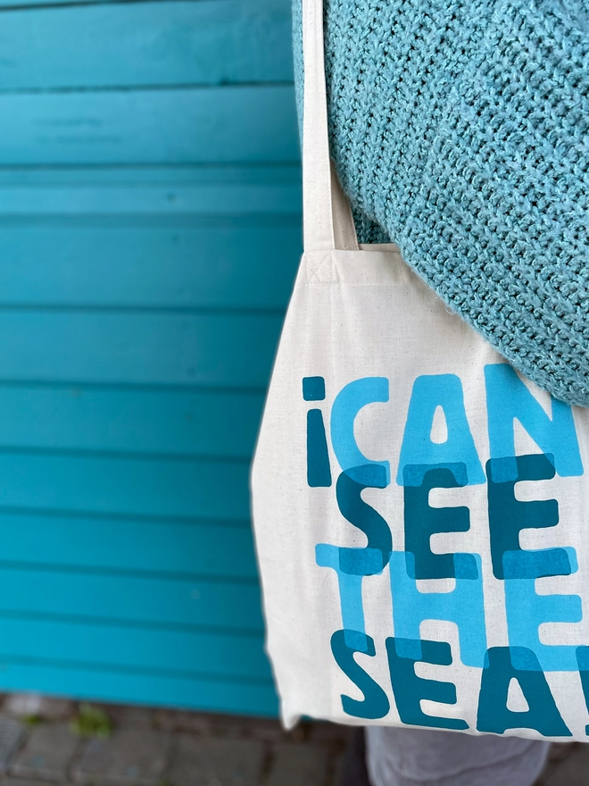  The words I Can See the Sea is screenprinted in two overlapping turquoise blues on this fairtrade cotton tote bag. Worn on the shoulder with a baggy cardigan sleeve showing, against a turquoise weather boarded background.
