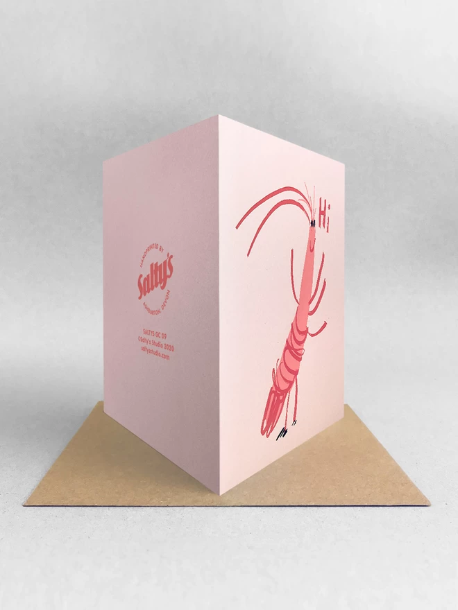 A happy cartoon prawn says Hi on this jolly upbeat pink card, viewed from behind showing front design and back brand stamp, stood on a Kraft envelope in a light grey studio space.