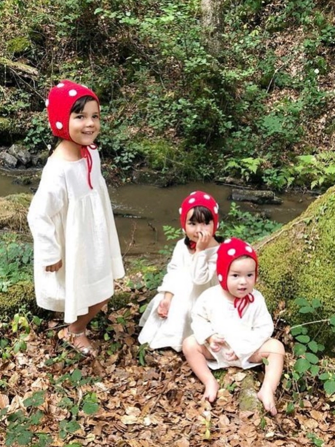 Three children in white nightgowns sit in woodland with red and white spotted bonnets on