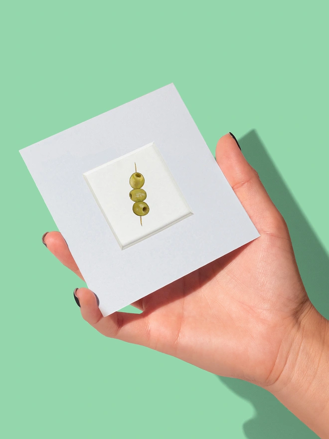 Hand holding a small print featuring three olives to show the size