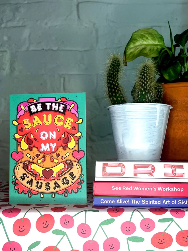 A green card featuring the phrase “Be the sauce on my sausage” on top of an abstract illustration including a hot dog, squirts of ketchup and mustard, and colourful hearts, sits on a shelf covered with illustrated cherry wrapping paper, next to books and two pot plants. 