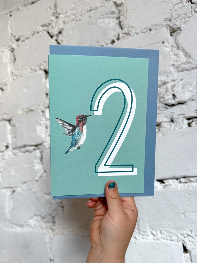 a pale teal card with a big white number 2 and an illustration of a hummingbird with a blue envelope
