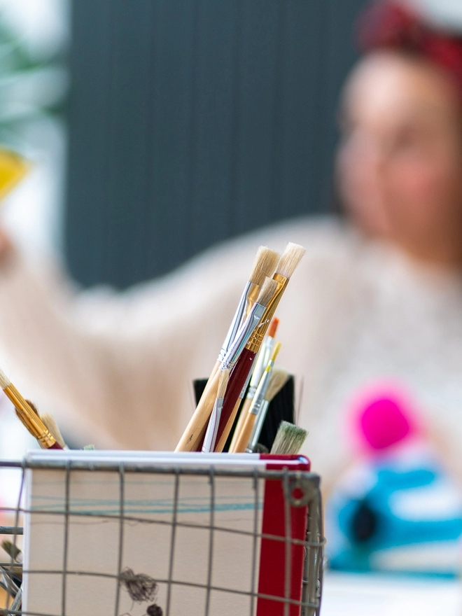Soft focused blurry image on Sketchy Muma in Studio behind some paintbrushes and a pink and blue teapot cosy