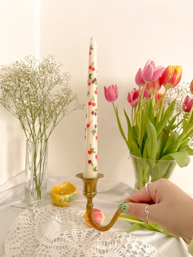 Mon Cherry & Tulip Fever Candle, a woman placing the candle on a table set accompanied by flowers in vases. It's floral design wraps around the stalk. 