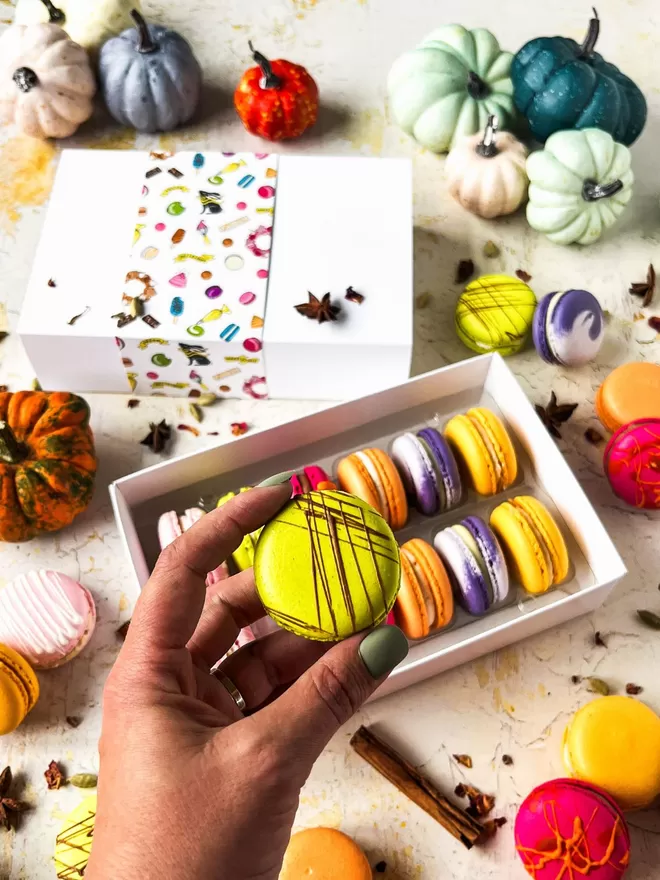 a person's hand holding a bright green macaron decorated with chocolate drizzle in front of a box of colourful autumn macarons surrounded by colourful pumpkins on a table 