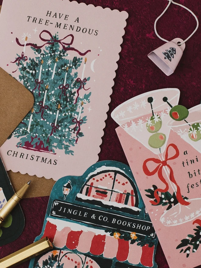 Bookstore Christmas card on a burgundy background with the Tree-mendous and Tini Bit Festive Christmas cards and a gold pen