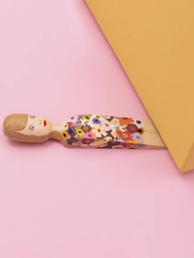 Wooden Door Stopper (Swifty Floral Outfit), a colourful wooden door stop sitting under a door.