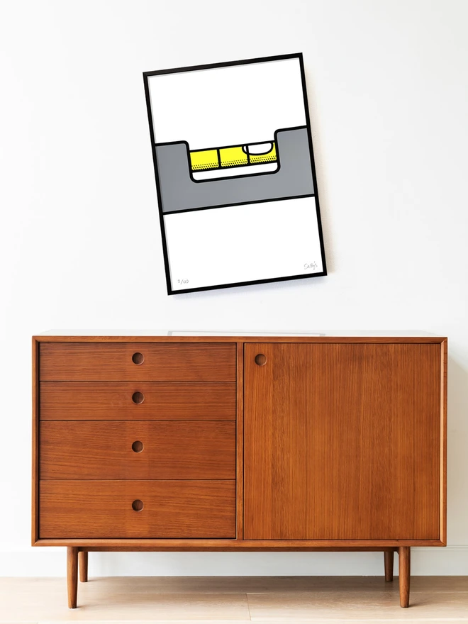 Screenprint of a spirit level with the bubble level, but the frame on the wonk. Above a mid century modern sideboard hung in a simple black frame on a white wall.