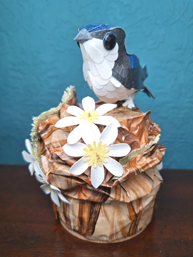 Housemartin bird sculpture, perched on a floral tree stump. This handmade sculpture is made from recycled paper. The bird is black and blue, with a white belly. White and yellow flowers adorn the tree stump. Front view.