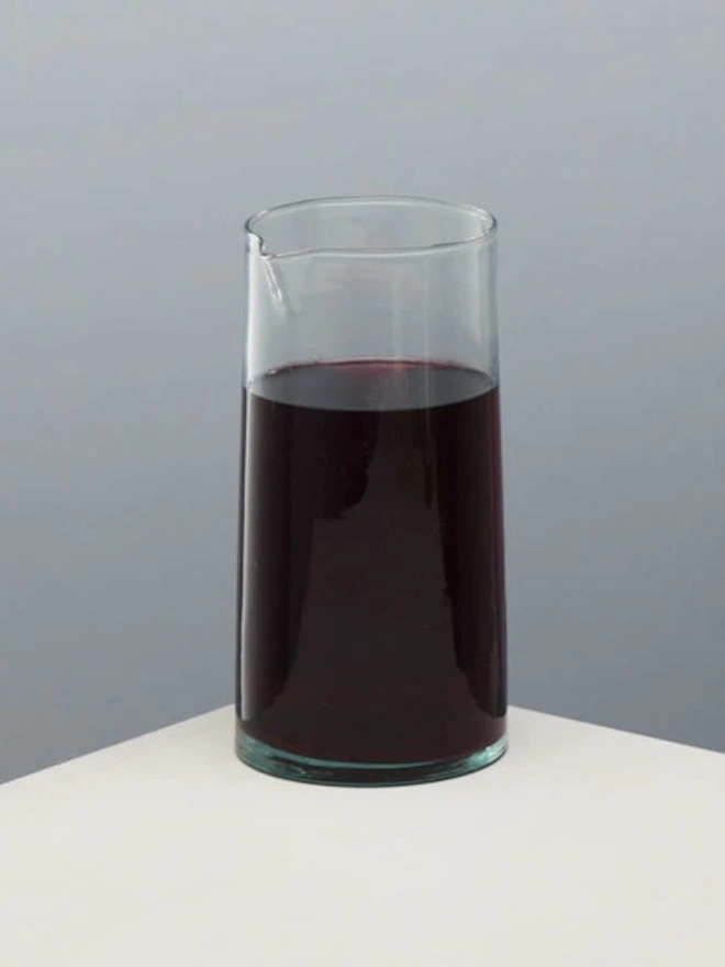 Glass Carafe, a large glass jug sitting on a plain counter top against a baby blue backdrop. It is filled by a wine. 