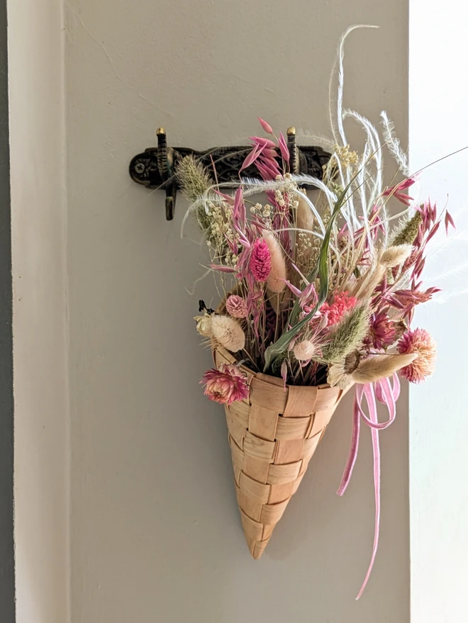 wooden braided cone basket with dried pink flowers hanging from a hook on a wall next to a green coat