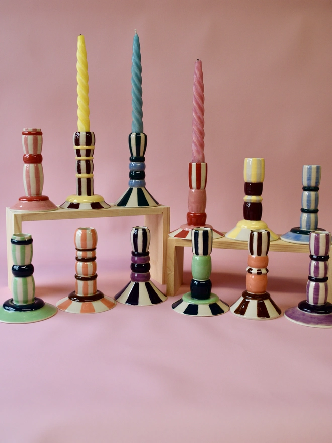 Collection of handmade ceramic candlesticks in multiple colourful stripe patterns, displayed on wooden stands with twisted candles, featuring pink, green, blue, yellow, and burgundy tones.