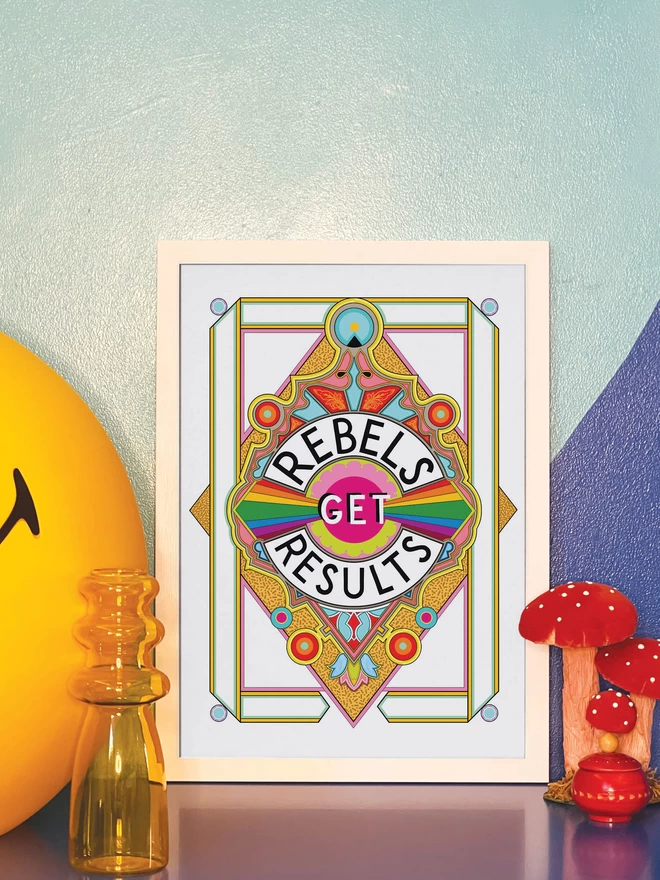 Rebels Get Results is written in black on a white background at the centre of this vibrant, abstract portrait illustration, with a white background and rainbows emitting from the centre, and multi-coloured detailing. The picture is in a white frame leaning against a painted blue wall on a blue cabinet. Next to the frame is a large illuminated yellow Smiley lamp, a yellow vase, a model of three red and white toadstools and a small round red wooden pot.