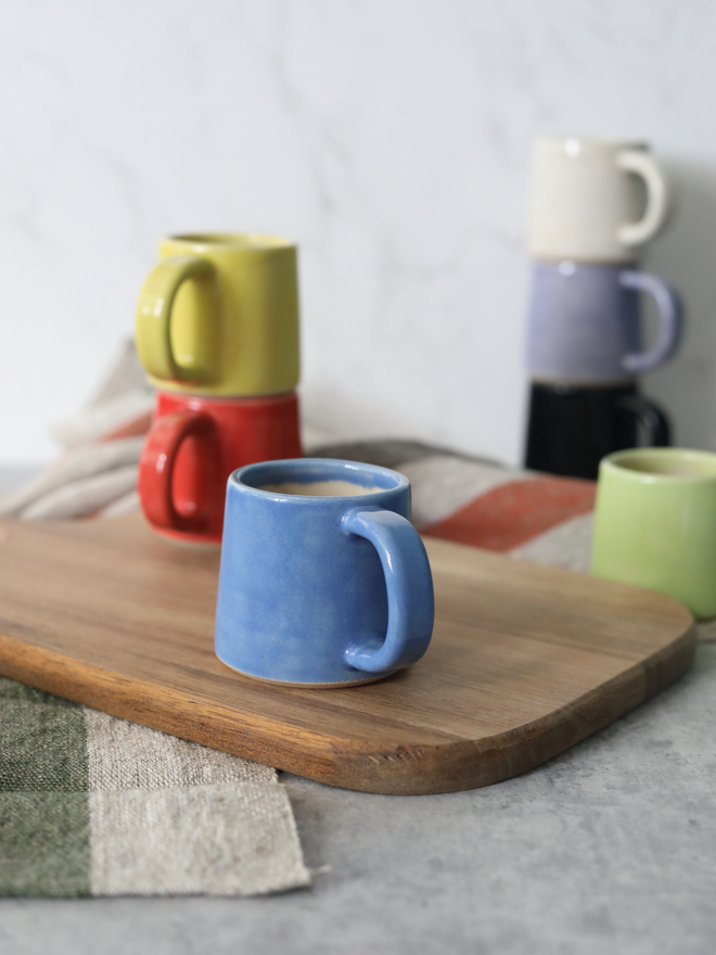 Several handmade ceramic espresso mugs arranged on a wooden board, with a pacific blue mug in the foreground holding espresso, and yellow, red, purple, black, white, and spring green mugs grouped behind, styled with linen cloths against a marble background