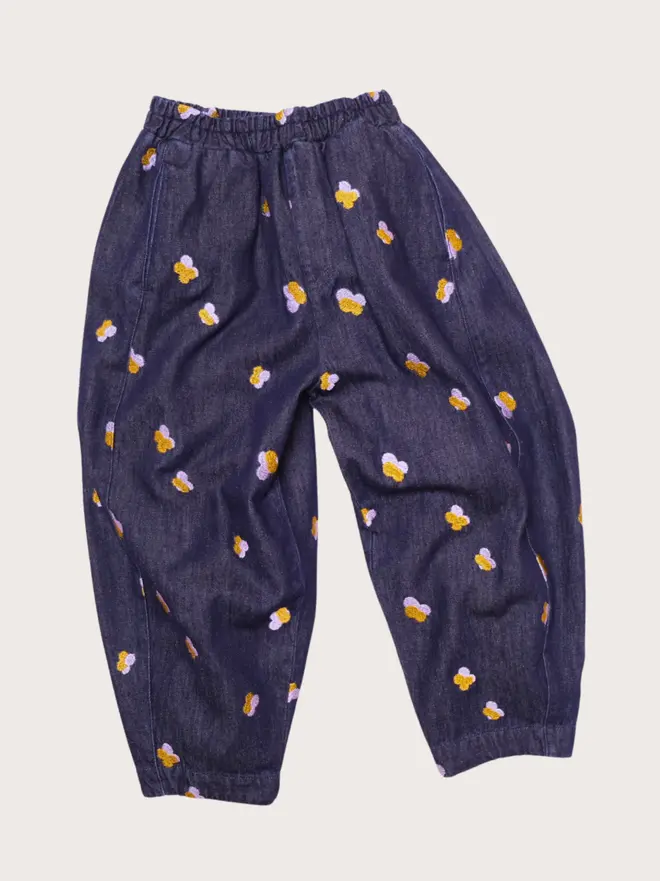 Front flat lay of Studio Koter Manu trousers in Denim Petal, dark denim barrel-leg trousers with elasticated waist and lilac and gold embroidered petals.