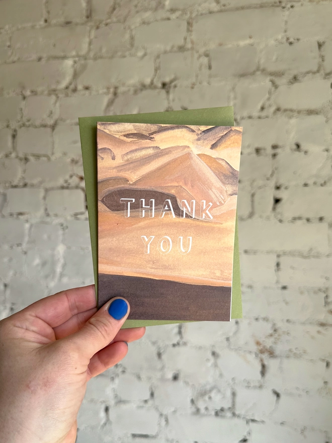 a greetings card featuring an illustration of a sand dunes landscape with the text “thank you” with a green envelope