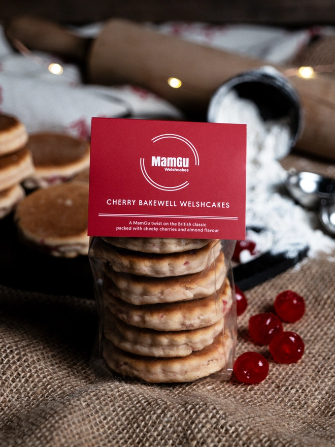 packaged cherry bakewell welsh cakes with a red MamGu Welshcakes Label on a busy kitchen worktop with spilt flour, rolling pin and cherries